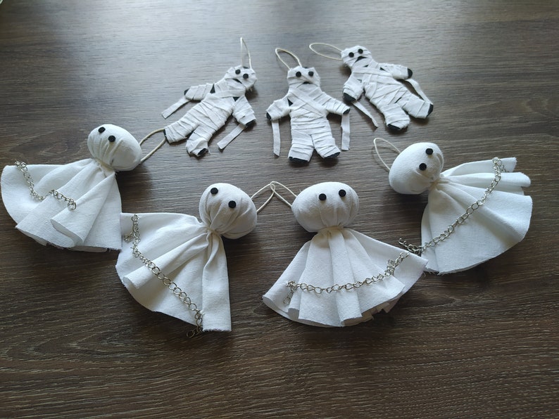 May include: A collection of seven handmade ornaments consisting of three white fabric mummy figures wrapped in strips and four white ghost figures with round heads, black bead eyes, and silver-toned metal chains draped across their fabric bodies.
