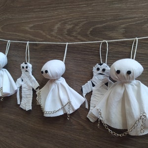 May include: A decorative garland featuring alternating white fabric ghost figures with black button eyes and mummy figures wrapped in white strips. Each ghost has a silver-toned metal chain draped across the front, and all figures hang from a white string.