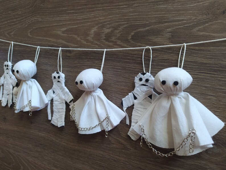 May include: A decorative garland featuring alternating white fabric ghost figures with black button eyes and mummy figures wrapped in white strips. Each ghost has a silver-toned metal chain draped across the front, and all figures hang from a white string.