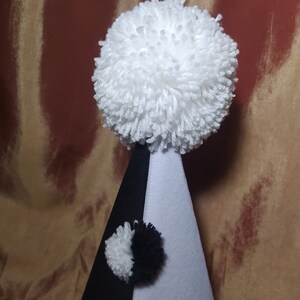 May include: A whimsical party hat in black, white, and gray. The hat is cone-shaped with a large white pom-pom at the top and a smaller pom-pom on the front. The hat is made of felt and yarn.