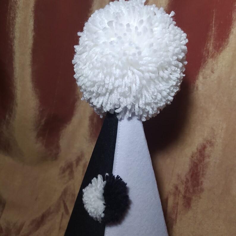 May include: A whimsical party hat in black, white, and gray. The hat is cone-shaped with a large white pom-pom at the top and a smaller pom-pom on the front. The hat is made of felt and yarn.