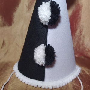 May include: A black and white felt party hat with two pom-poms. The hat is divided in half, with one side black and the other white. The base is trimmed with white pom-pom trim and has a white string.