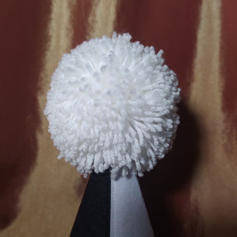 May include: A close-up of a white pom-pom atop a black and white cone-shaped object. The pom-pom is made of fluffy yarn. The background is a mix of brown and gold vertical stripes.