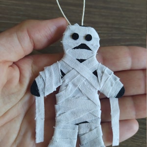 May include: A small mummy ornament crafted from black felt wrapped in strips of white fabric. The figure features two circular black felt eyes and a white string loop at the top for hanging. The mummy has dangling fabric strips at the arms.