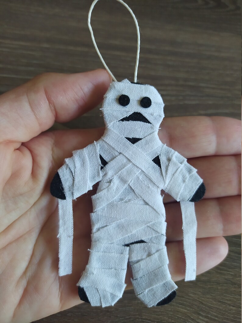 May include: A small mummy ornament crafted from black felt wrapped in strips of white fabric. The figure features two circular black felt eyes and a white string loop at the top for hanging. The mummy has dangling fabric strips at the arms.