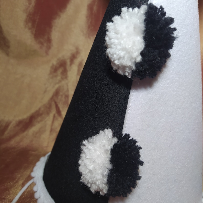 May include: A black and white felt party hat with two pom-poms. The hat is divided diagonally, with one side black and the other white. The pom-poms are half black and half white. The hat has a white scalloped trim.
