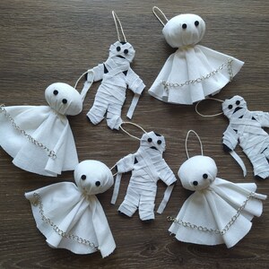 May include: A collection of six handmade ornaments featuring three white fabric ghost figures with silver chains and three white fabric mummy figures wrapped in strips of cloth, each with black button eyes and a hanging loop for display.