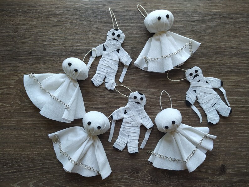 May include: A collection of six handmade ornaments featuring three white fabric ghost figures with silver chains and three white fabric mummy figures wrapped in strips of cloth, each with black button eyes and a hanging loop for display.