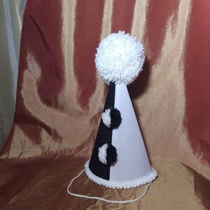May include: A party hat with a black and white color scheme. The hat is divided vertically, with one half black and the other white. It features a large white pom-pom on top and smaller black and white pom-poms. A white string is attached.