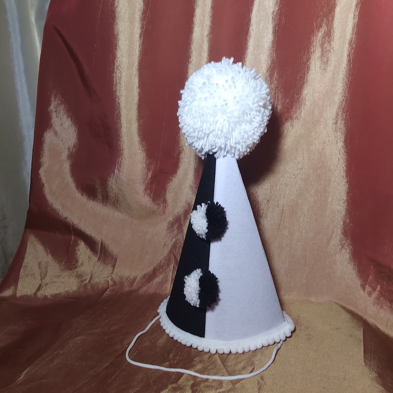 May include: A party hat with a black and white color scheme. The hat is divided vertically, with one half black and the other white. It features a large white pom-pom on top and smaller black and white pom-poms. A white string is attached.