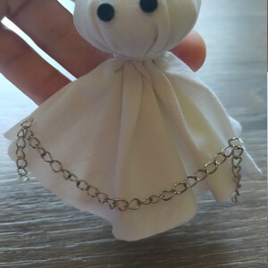 May include: A small, white fabric doll with a rounded head and two black circular eyes. The doll features a draped, skirt-like body made of white cloth, accented by a thin, metallic silver-toned chain draped across the front of the fabric.