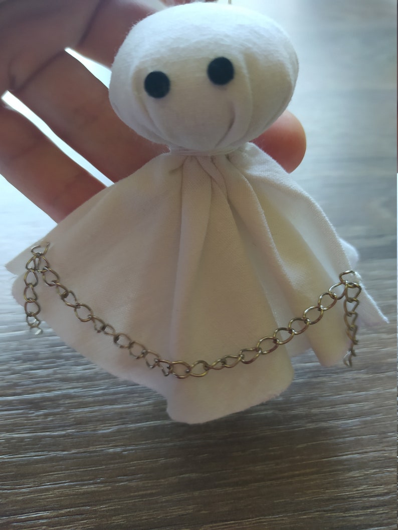 May include: A small, white fabric doll with a rounded head and two black circular eyes. The doll features a draped, skirt-like body made of white cloth, accented by a thin, metallic silver-toned chain draped across the front of the fabric.