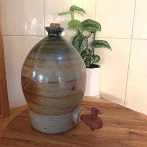 May include: A large, grey, brown and white ceramic jug with a wooden cork stopper and a metal spigot. The jug is sitting on a wooden surface.