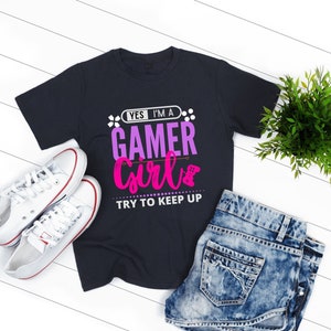 May include: A dark blue t-shirt with the text "Yes I'm a Gamer Girl Try to Keep Up" in pink and purple lettering. The shirt is on a white wooden surface with a pair of white trainers and denim shorts.