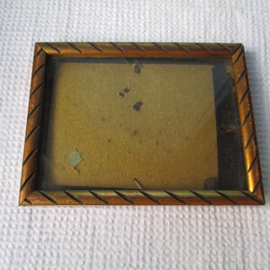 Könnte beinhalten: Ein goldfarbener Bilderrahmen mit Holzmaserung und abgeschrägter Kante. Der Rahmen hat eine Glasfront und eine braune Papierrückseite.