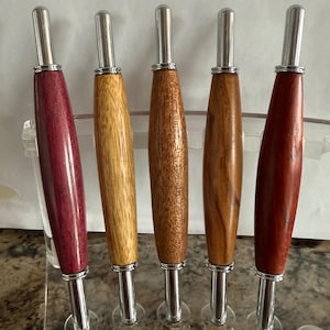 May include: Five wooden bottle stoppers with silver metal accents. The stoppers are displayed in a clear acrylic holder. The wood colors vary from light to dark brown and a deep reddish-purple.