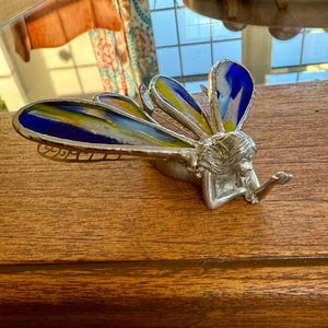 May include: A silver fairy figurine with colorful stained-glass wings in shades of blue, yellow, and green. The fairy is lying down, with one arm extended. The wings have a detailed, textured design.
