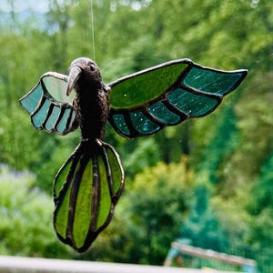 May include: A stained glass hummingbird suncatcher featuring a dark brown metal body and wings with translucent green and teal glass panels. The bird is suspended by a thin wire, displaying segmented wing and tail feathers in a flying pose.