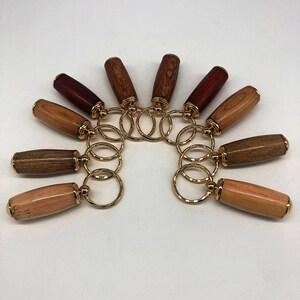 May include: A collection of wooden keychains with gold-colored metal accents. The keychains feature various wood tones, including light brown, dark brown, and reddish-brown. Each keychain has a cylindrical wooden body and a gold-colored ring.