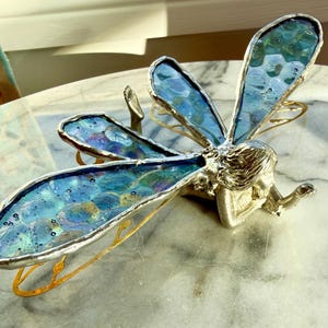 May include: A decorative dragonfly sculpture with iridescent blue and green glass wings. The wings are outlined in silver metal, and the body is silver-toned. The dragonfly rests on a marble surface, with gold-colored accents.