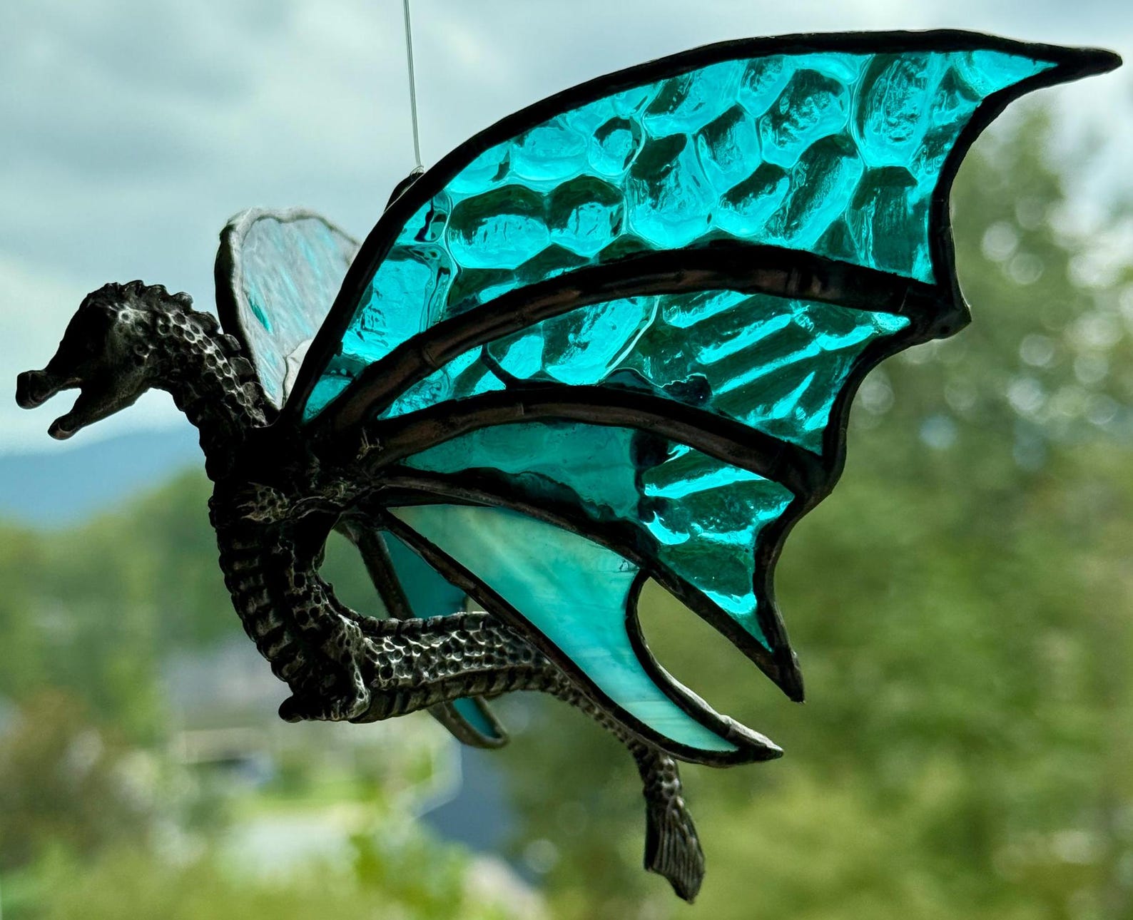 A metal dragon sculpture with aqua colored wings hangs in a window.