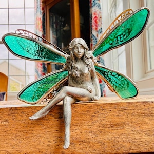 May include: A silver-toned fairy figurine with large, green-tinted wings. The fairy is seated with one leg crossed over the other. The wings have a decorative metal frame. The figurine is placed on a wooden surface.