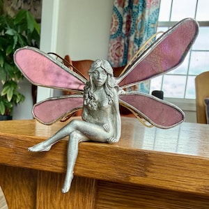 May include: A silver-colored fairy figurine with pink and gold-trimmed wings. The fairy is seated with one leg extended. The wings are a translucent pink. The figurine is on a wooden surface.