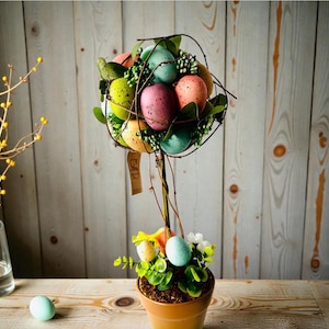 May include: An Easter topiary arrangement featuring speckled eggs in various colours. The eggs are held within a wire frame, with foliage and small white flowers at the base. The terracotta pot sits on a wooden surface, with a single egg nearby.