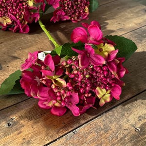 May include: Close-up of vibrant pink artificial hydrangea flowers with green leaves. The flowers are clustered together, showcasing a mix of textures and shades of pink and yellow. The arrangement rests on a rustic wooden surface.