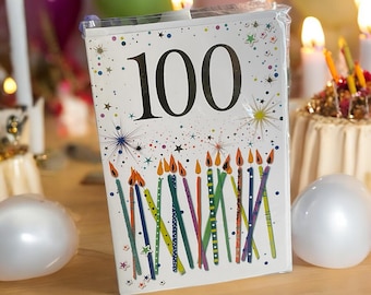 May include: A birthday card with the number 100 in large black font. The card is decorated with colorful candles and starbursts. Lit candles and balloons are in the background, creating a festive atmosphere.