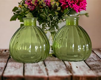 May include: Two green glass vases with a ribbed texture. The vases are round and have a wide mouth. They are sitting on a wooden table.