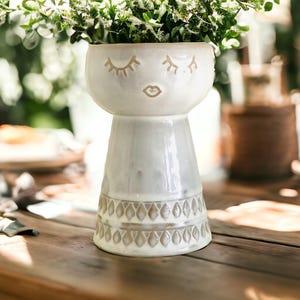 May include: White ceramic vase with a face design featuring closed eyes and a smiling mouth. The vase has a textured base with a geometric pattern.