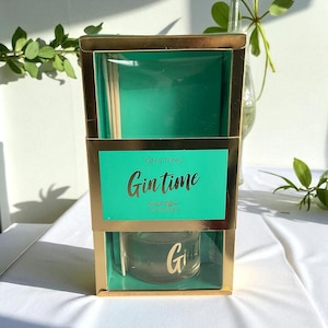 Op de afbeelding: Een goudomrand cadeaupakket met een turquoise interieur bevat een glazen fles, houten stokjes en een turquoise kaart. De kaart toont "Gin & Tonic, Gin Time, Scented Diffuser, Candlelight" in gouden letters. De fles heeft een gouden "G".