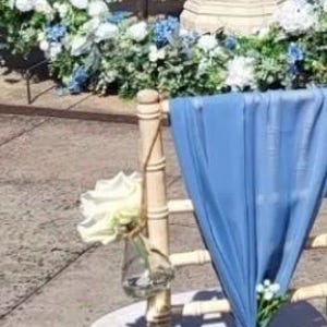 May include: A wedding ceremony setup featuring a decorated chair with a light blue fabric drape and floral accents. A small glass vase holds a white rose. The background includes a floral arch with white and blue flowers.