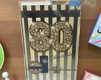 Puede incluir: Tarjeta de cumpleaños con el número 80 dorado, adornado con gemas brillantes, sobre un fondo a rayas. La tarjeta incluye las frases "Happy Birthday" y "Celebrate Today!" con elementos decorativos.