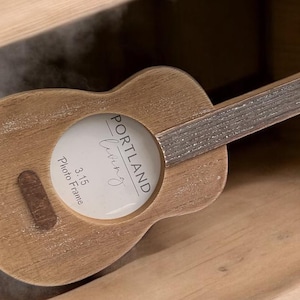 May include: A wooden guitar-shaped photo frame with a 3.15 inch opening. The frame is painted brown and has a distressed finish. The text "PORTLAND" is printed on the frame.