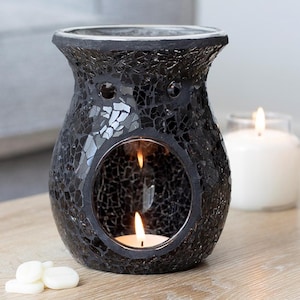 May include: Black mosaic glass oil warmer with a round opening and a lit tea light candle inside. The warmer has a wide base and a flared top. White wax melts are in the foreground. A lit white candle is in the background.