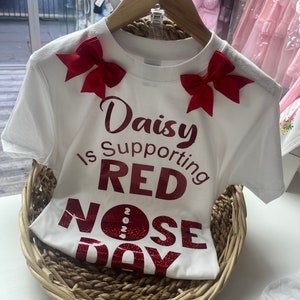 May include: White t-shirt with red bows and text that reads "Daisy Is Supporting RED NOSE DAY 2024". The shirt is displayed in a woven basket, suggesting a children's clothing item.