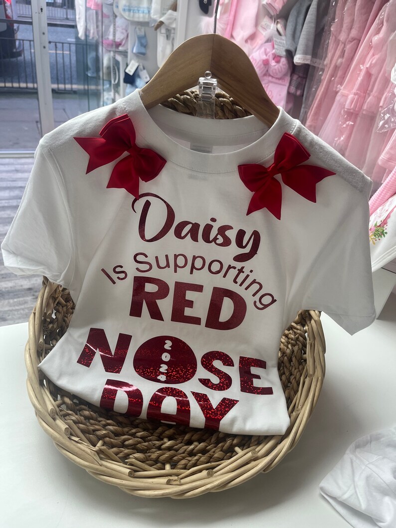 May include: White t-shirt with red bows and text that reads "Daisy Is Supporting RED NOSE DAY 2024". The shirt is displayed in a woven basket, suggesting a children's clothing item.