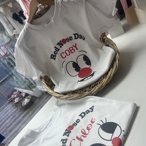 May include: Two white t-shirts with a cartoon face design for Red Nose Day. One shirt says "Red Nose Day COBY" and the other says "Red Nose Day Chloe". The shirts are in a wicker basket and on a white surface.