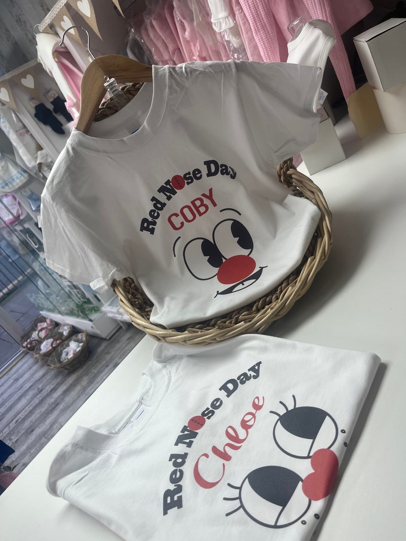 May include: Two white t-shirts with a cartoon face design for Red Nose Day. One shirt says "Red Nose Day COBY" and the other says "Red Nose Day Chloe". The shirts are in a wicker basket and on a white surface.