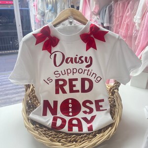May include: White t-shirt with red bows on the shoulders and the text "Daisy is Supporting RED NOSE DAY 2024" in red glitter. The shirt is displayed in a woven basket, suggesting a children's clothing item.