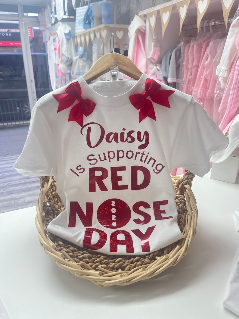 May include: White t-shirt with red bows on the shoulders and the text "Daisy is Supporting RED NOSE DAY 2024" in red glitter. The shirt is displayed in a woven basket, suggesting a children's clothing item.