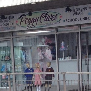 May include: A storefront for Peggy Clives, a children's wear and school uniform shop. The sign displays the shop's name in a cursive font, along with services like personalized printing and embroidery. Mannequins in school uniforms and dresses are visible in the window.
