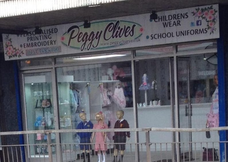 May include: A storefront for Peggy Clives, a children's wear and school uniform shop. The sign displays the shop's name in a cursive font, along with services like personalized printing and embroidery. Mannequins in school uniforms and dresses are visible in the window.