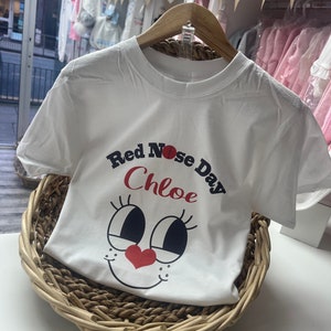 May include: White t-shirt with the text "Red Nose Day Chloe" and a cartoon face design. The shirt is displayed on a wooden hanger inside a woven basket. The design features a red nose and heart.