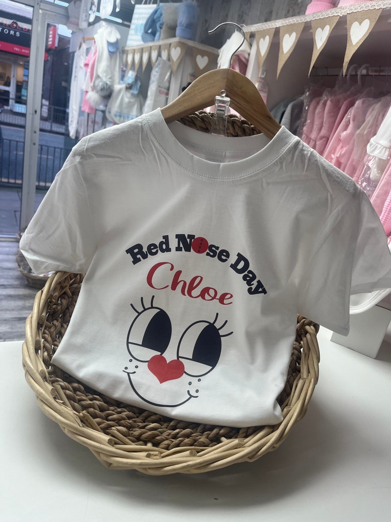 May include: White t-shirt with the text "Red Nose Day Chloe" and a cartoon face design. The shirt is displayed on a wooden hanger inside a woven basket. The design features a red nose and heart.