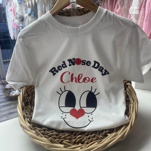 May include: White t-shirt with a graphic design for Red Nose Day, featuring a cartoon face with large eyes, a red nose, and a heart-shaped nose. The name "Chloe" is printed above the face. The shirt is displayed in a woven basket.