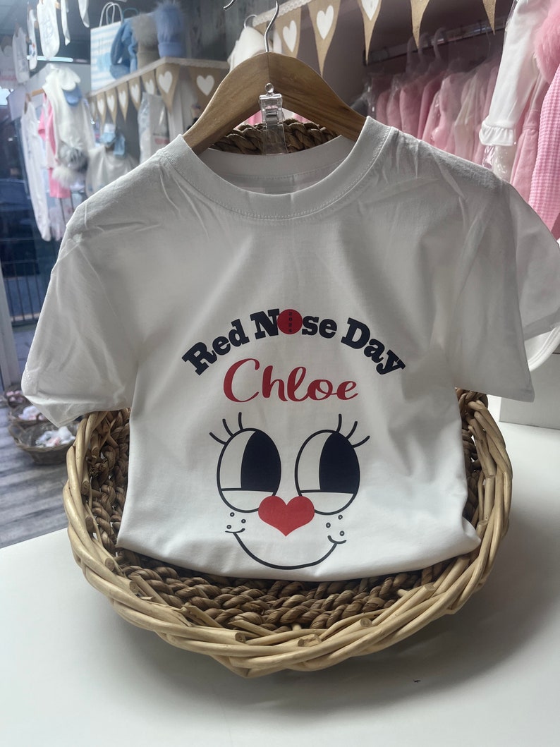 May include: White t-shirt with a graphic design for Red Nose Day, featuring a cartoon face with large eyes, a red nose, and a heart-shaped nose. The name "Chloe" is printed above the face. The shirt is displayed in a woven basket.