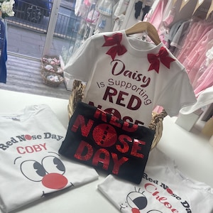 May include: White and black t-shirts with Red Nose Day graphics. One shirt has the text "Daisy is Supporting RED NOSE DAY" with red bows. Other shirts feature cartoon faces and names like "COBY" and "Chloe".